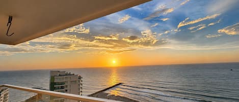Une vue sur le coucher de soleil depuis un balcon donnant sur une plage, avec une jetée et des bâtiments au premier plan.