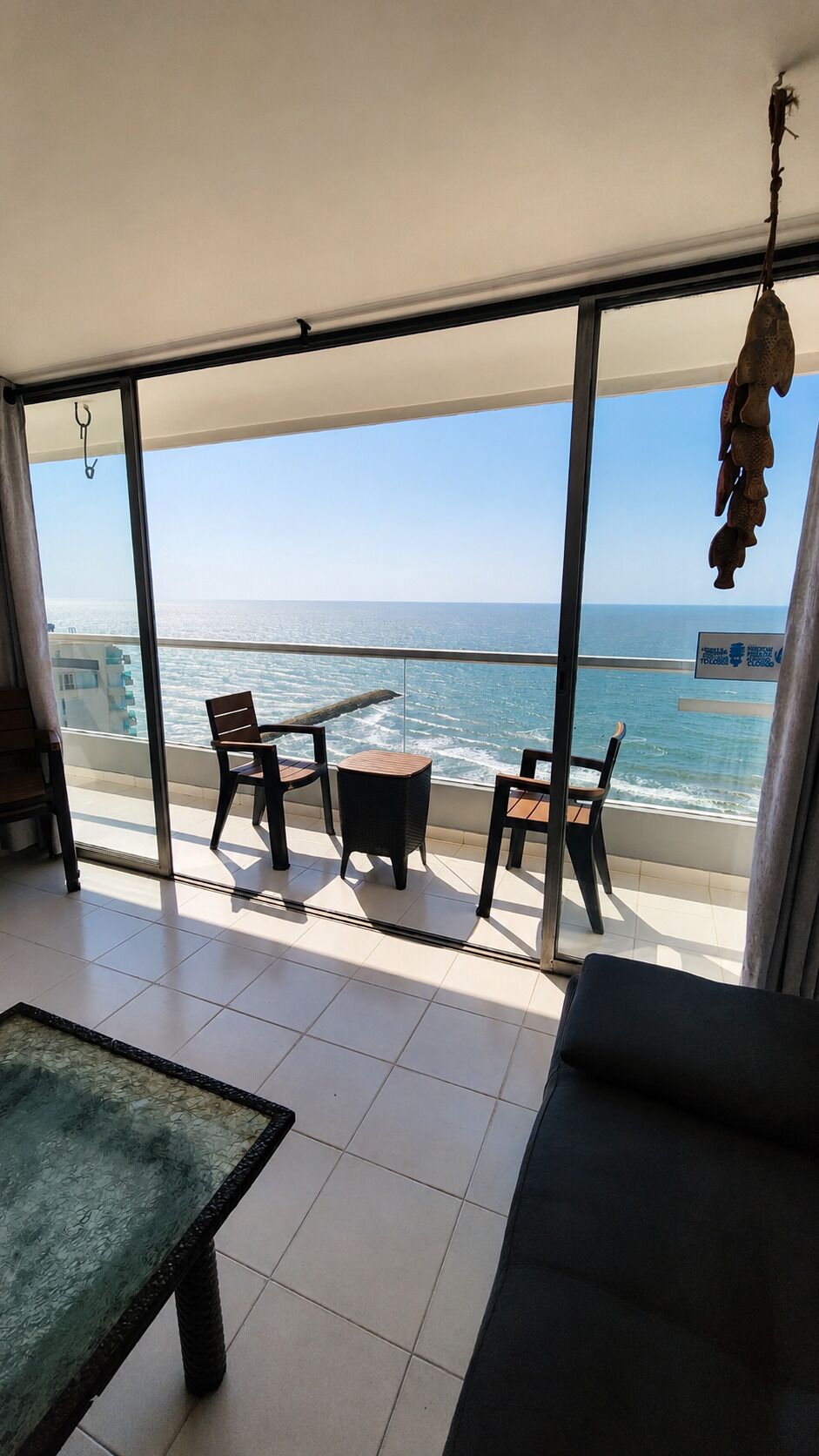 A balcony with a view of the sea, furnished with a table and chairs, and a hanging decoration.