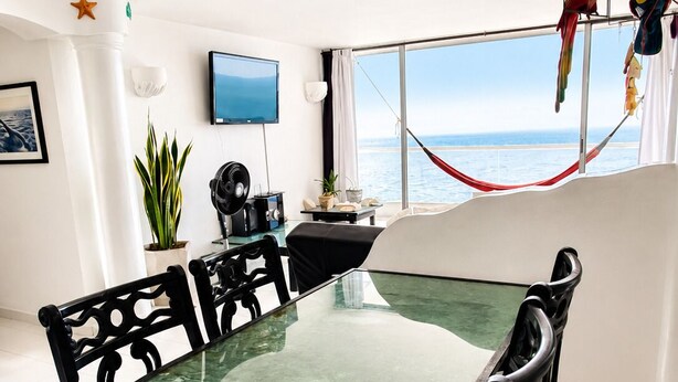 A dining area with a glass-top table, black chairs, a television, a plant, and a view of the sea through a window with a red hammock. - Ocean view with balcony—pool—Bocagrande Beach—Pet-friendly (Cartagena de Indias)