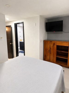 Une chambre à coucher comprenant un lit, une armoire en bois, un téléviseur fixé au mur et une porte donnant accès à une salle de bain.