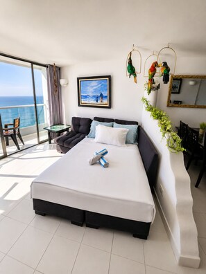 A hotel room with a large bed, a balcony with a view of the ocean, a dining area, and a living space.