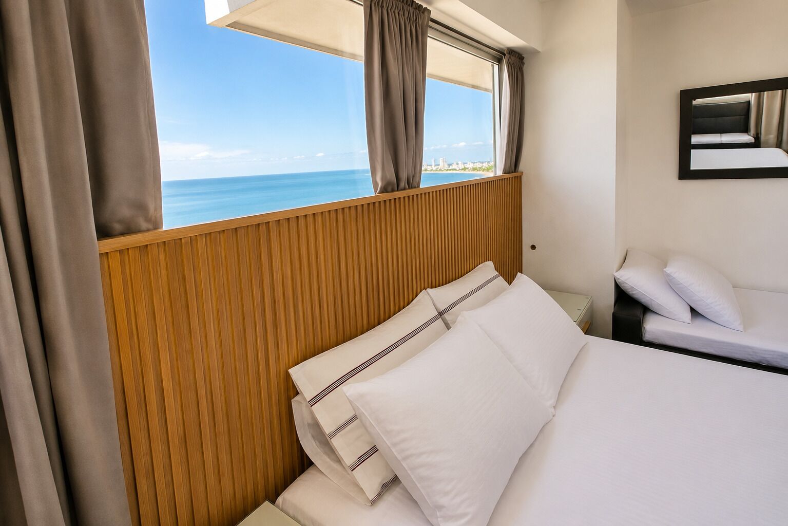 Ein Hotelzimmer mit Bett, Nachttisch, einem Fenster mit Vorhängen und Meerblick.