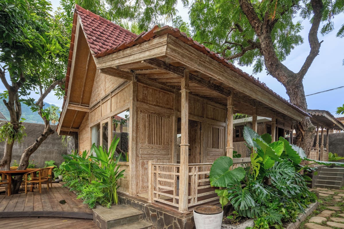 A wooden house with a tiled roof, surrounded by lush greenery and large trees.
