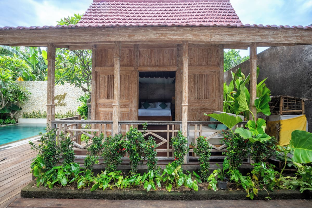 A wooden gazebo with a tiled roof, surrounded by lush greenery and a pool.