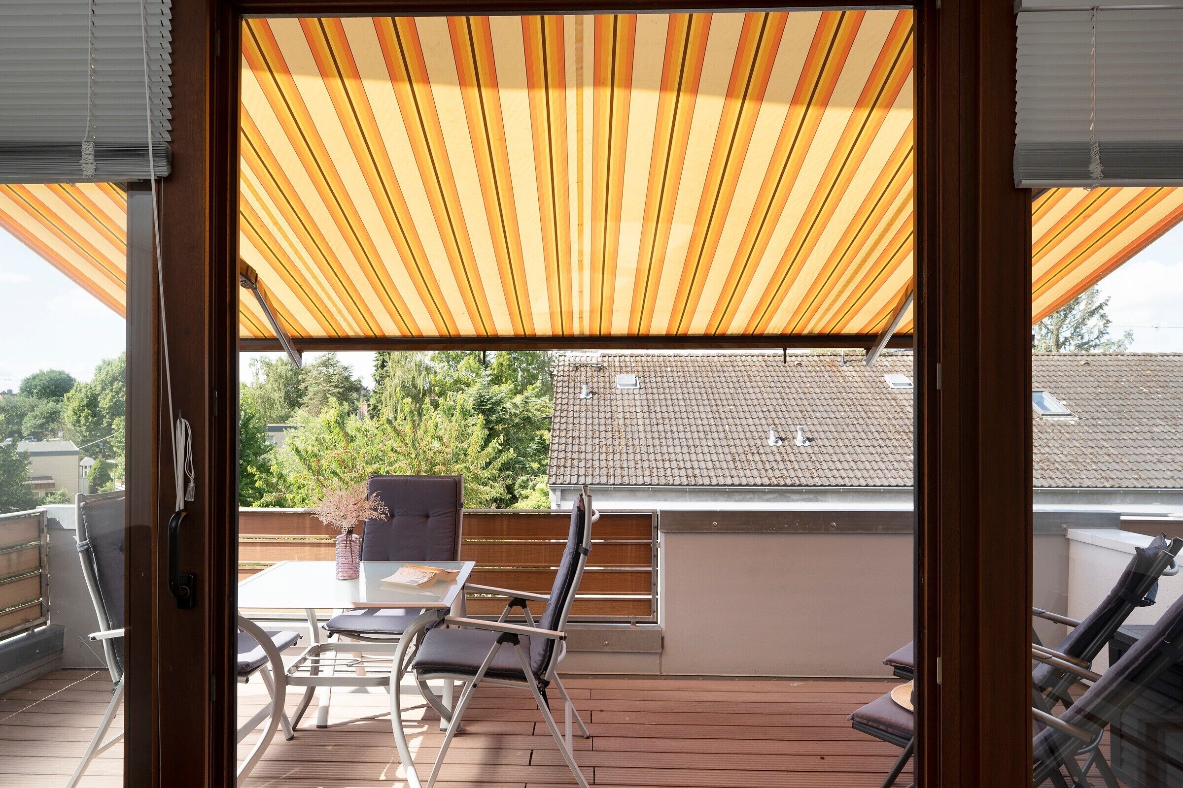 A balcony with a striped awning, a table, chairs, and a view of the neighborhood.