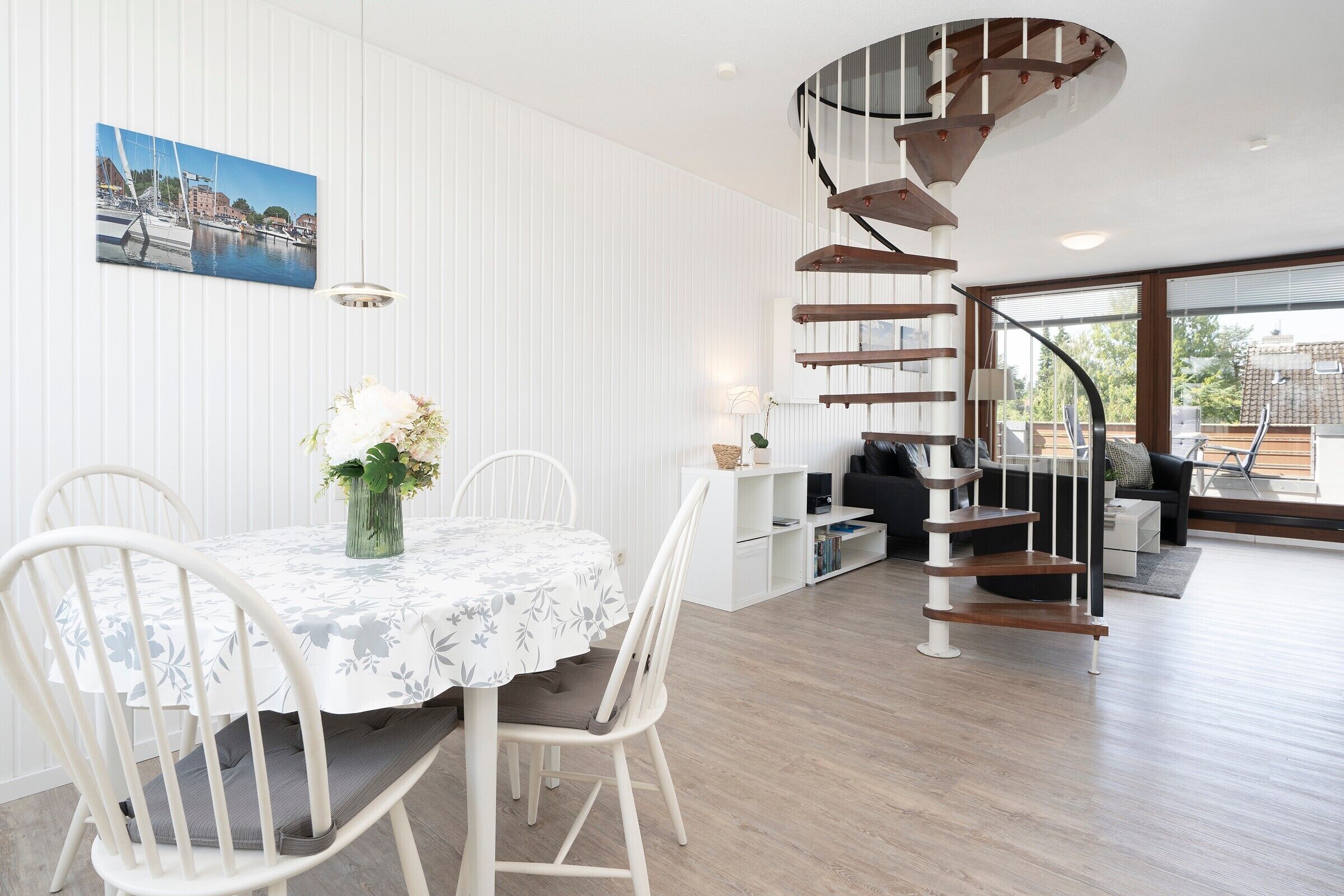 A dining area with a white table and chairs, a spiral staircase, and a cityscape painting on the wall.