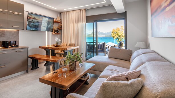 A modern living room with a sofa, wooden coffee table, and a dining area with a table and chairs. A large window offers a view of the sea and mountains. - Seaside Suite 3-Modern & Comfort (Episkopos)