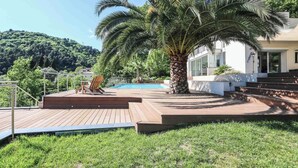 A wooden deck with a palm tree, a pool, and a white building with steps leading up to it.