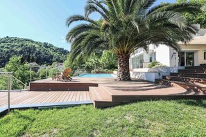 Une terrasse en bois avec un palmier, une piscine et un bâtiment blanc avec des marches menant à l’entrée.