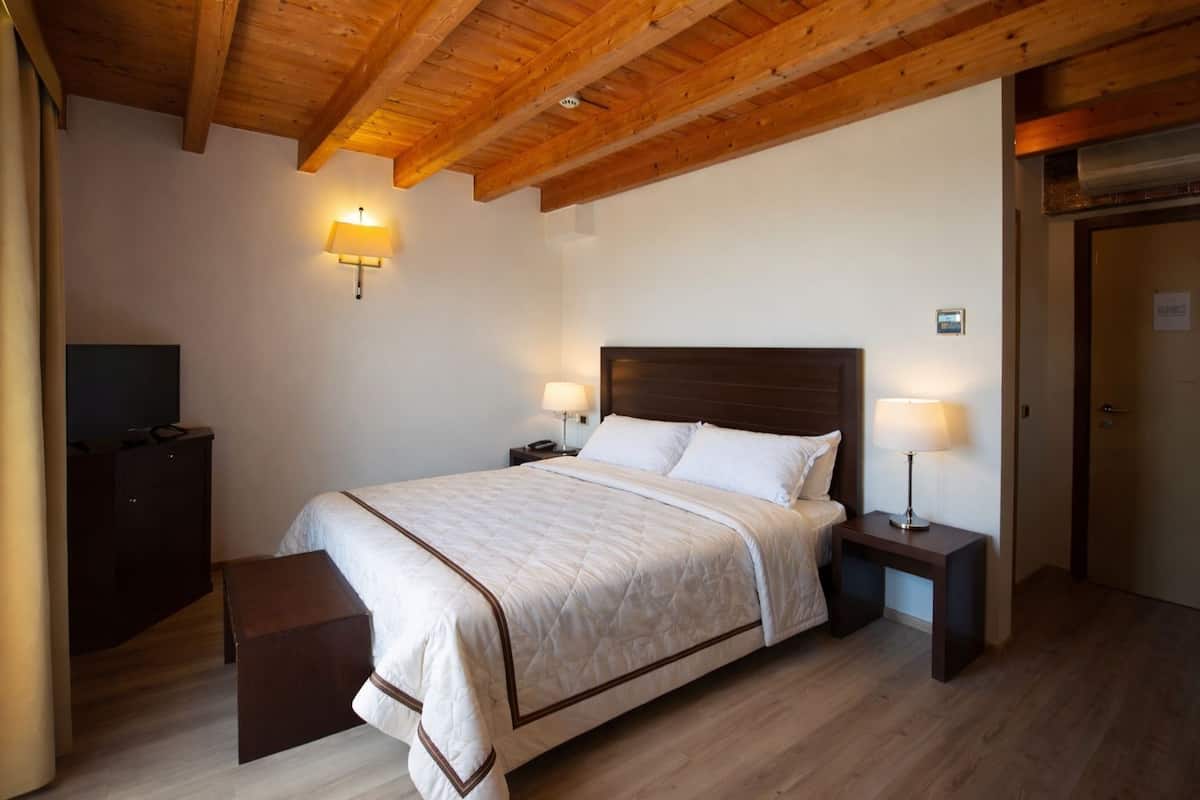 Una habitació amb sostre de fusta, un llit amb llençol blanc, dues taules de nit amb làmpades, un televisor sobre un moble i una porta.