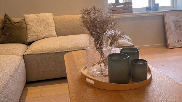 Ein Wohnzimmer mit einem beige gefärbten Sofa, einem Couchtisch aus Holz mit Vasen und Kerzen sowie einem Fenster mit Blick auf Gebäude.