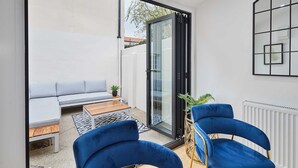 A modern patio with a sliding glass door, a grey sofa, a wooden table, and two blue chairs. - Host Stay Ginger Bread Cottage (Middlesbrough)