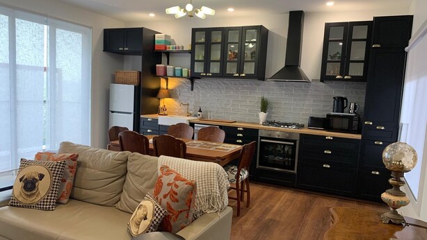 A modern kitchen with dark cabinets, a central island, and a dining area with a sofa and chairs. - La Maisonnette (Daylesford)