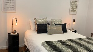 A neatly arranged bedroom with a large bed, two bedside tables with lamps, a black trash bin, and patterned wall art. - La Maisonnette (Daylesford)