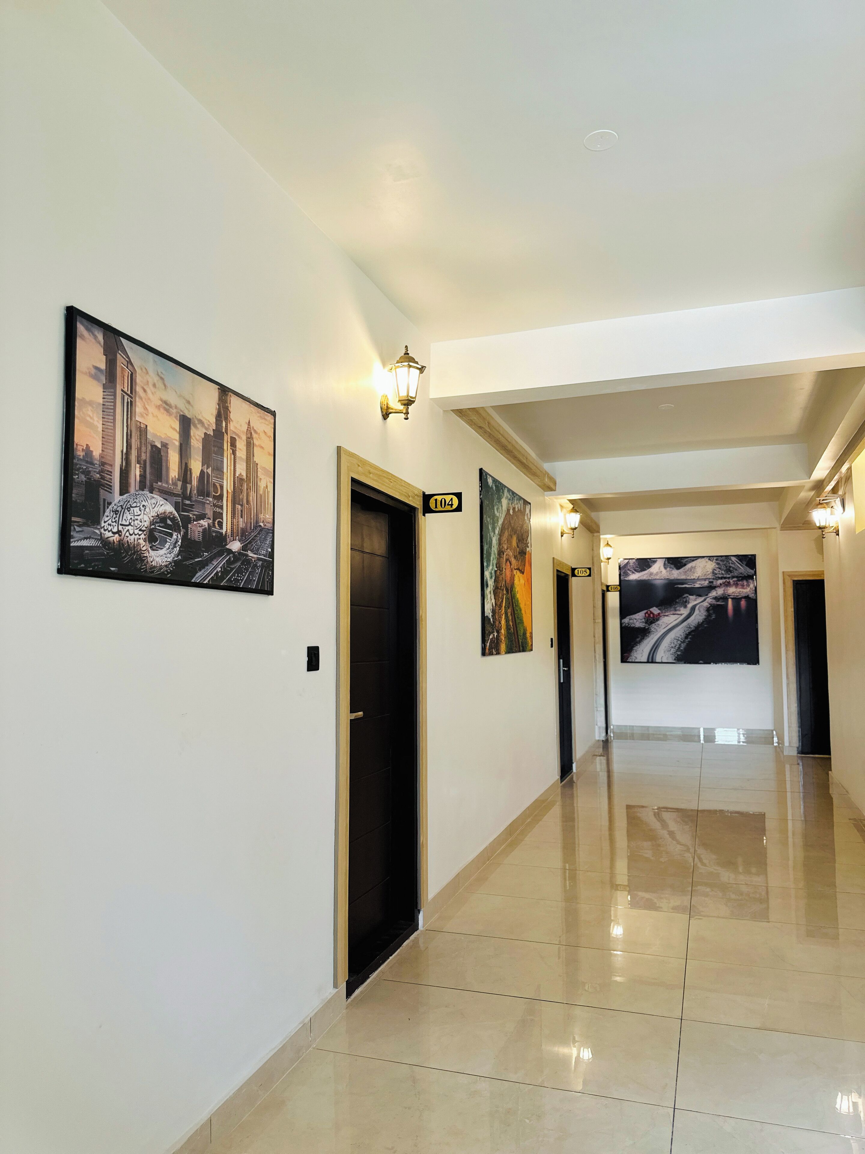 A hotel hallway with a modern design, featuring a large cityscape painting, a wooden door labeled "104", and a large landscape painting on the wall.