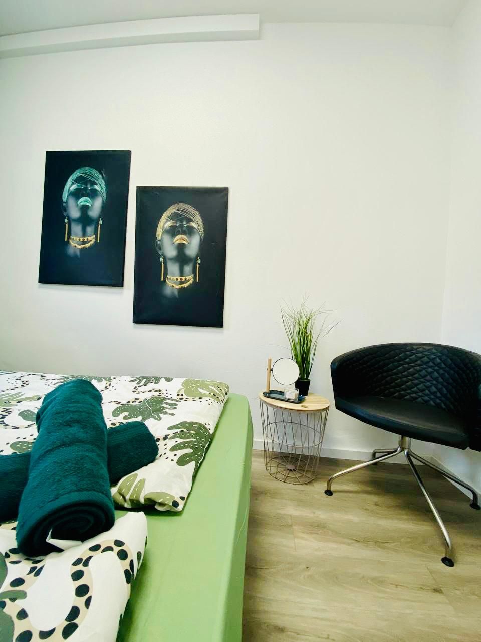 Ein Schlafzimmer mit einem Bett, einem Sessel, einem kleinen runden Tisch mit einer Pflanze und zwei an der Wand befestigten Kunstwerken.