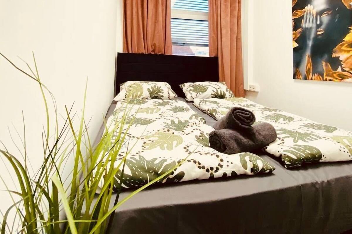 Un dormitorio con cama, cortinas, una ventana, un cuadro y una planta.
