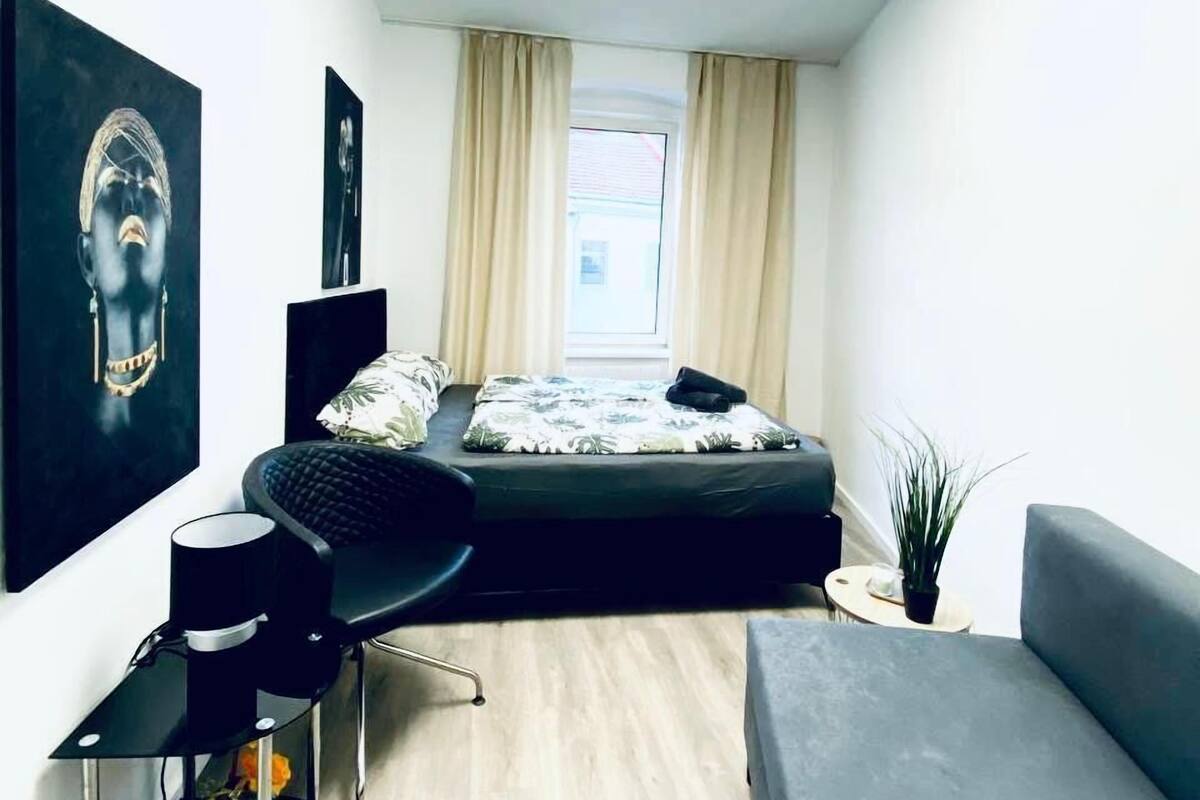 Un dormitorio moderno con una cama, un escritorio y una silla. Hay dos cuadros grandes en la pared, una ventana con cortinas y una planta en el suelo.