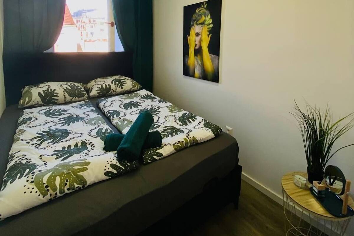 Un dormitorio con cama, ventana con cortinas, un cuadro en la pared y una pequeña mesa con una planta.