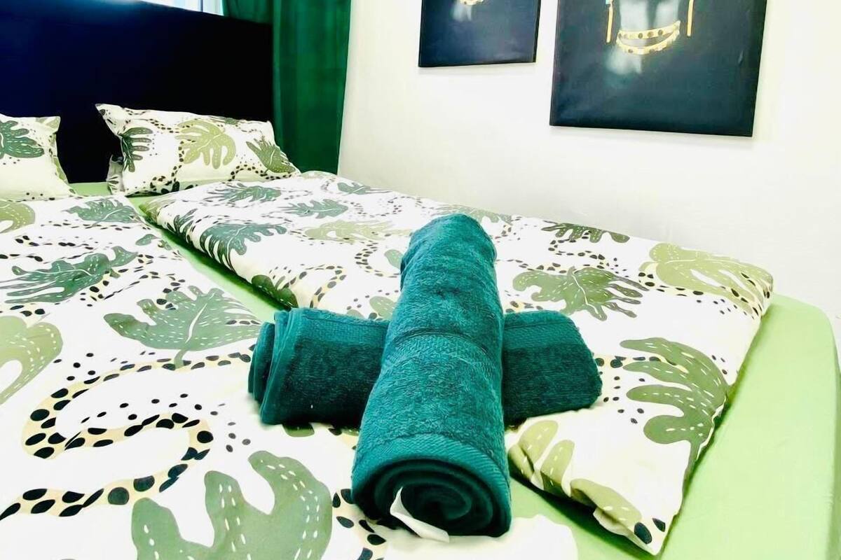 Una cama bien hecha con una manta verde y una toalla enrollada, una ventana con cortinas y dos cuadros en la pared.