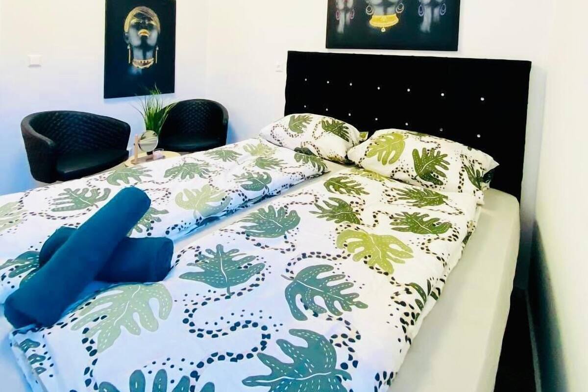 Un dormitorio con una cama, dos sillas negras, una pequeña planta en maceta y obras de arte en las paredes.
