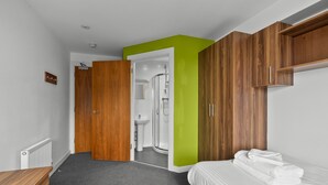 A modern hotel room with a wooden wardrobe, a green accent wall, a white bed with towels, and a bathroom with a shower and sink. - 8 Bed Apartment @ Derriford Stays by Pureserviced (Plymouth)