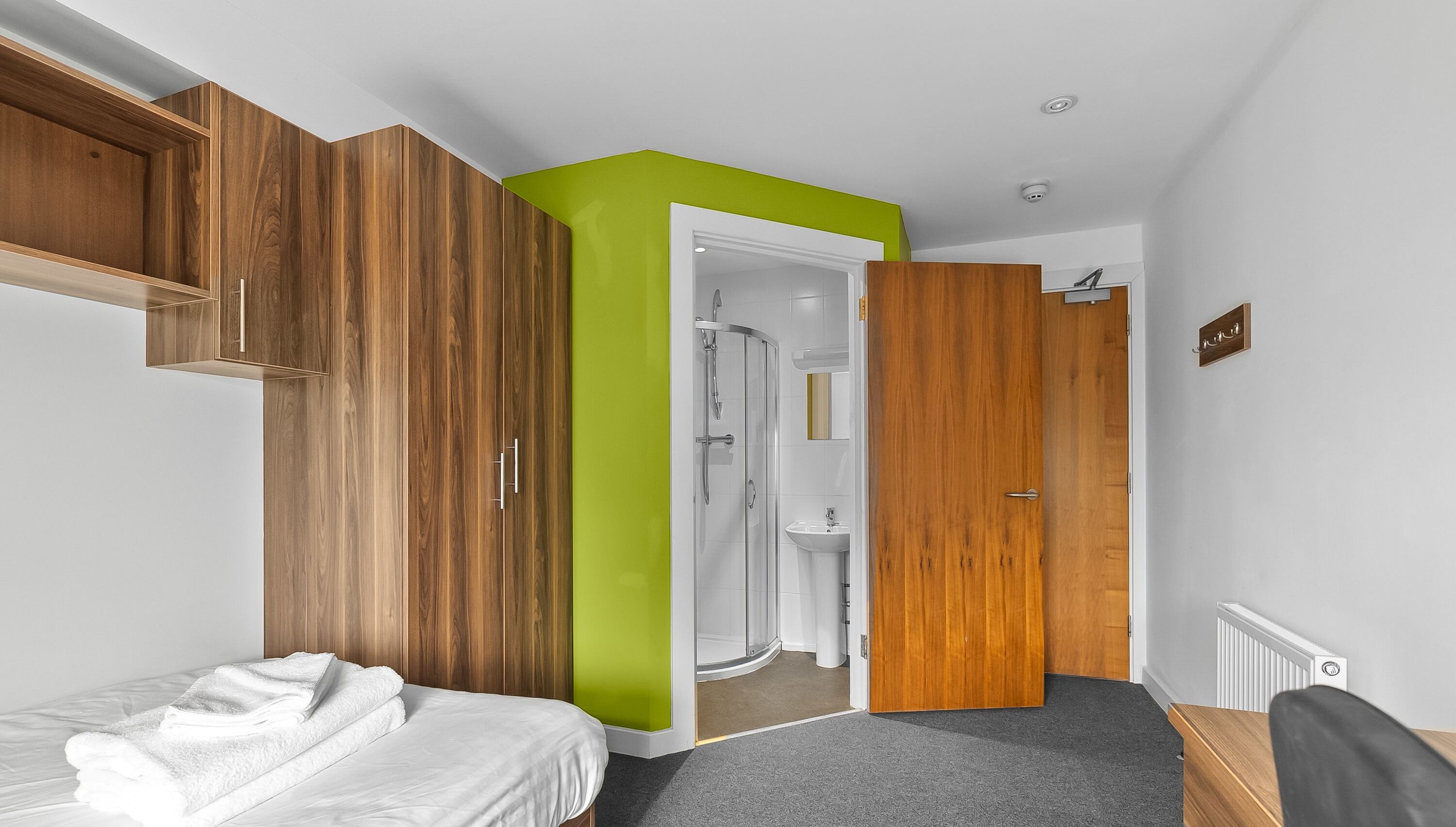 A hotel room with a bed, a wooden wardrobe, a green accent wall, a shower area, and a radiator.
