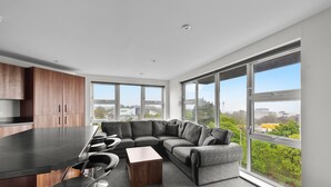 A modern kitchen with a black countertop and wooden cabinets, a gray sectional sofa with a wooden coffee table, and large windows offering a view of greenery and buildings. - 8 Bed Apartment @ Derriford Stays by Pureserviced (Plymouth)