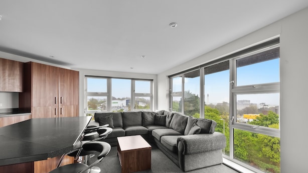 A modern kitchen with a black countertop and wooden cabinets, a gray sectional sofa with a wooden coffee table, and large windows offering a view of greenery and buildings. - 5 Bed Apartment @ Derriford Stays by Pureserviced (Plymouth)