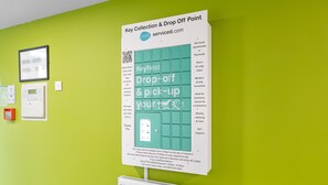 A Key Collection & Drop Off Point sign with a QR code, a framed architectural drawing, and a PLYM10 device on a green wall. - 5 Bed Apartment @ Derriford Stays by Pureserviced (Plymouth)