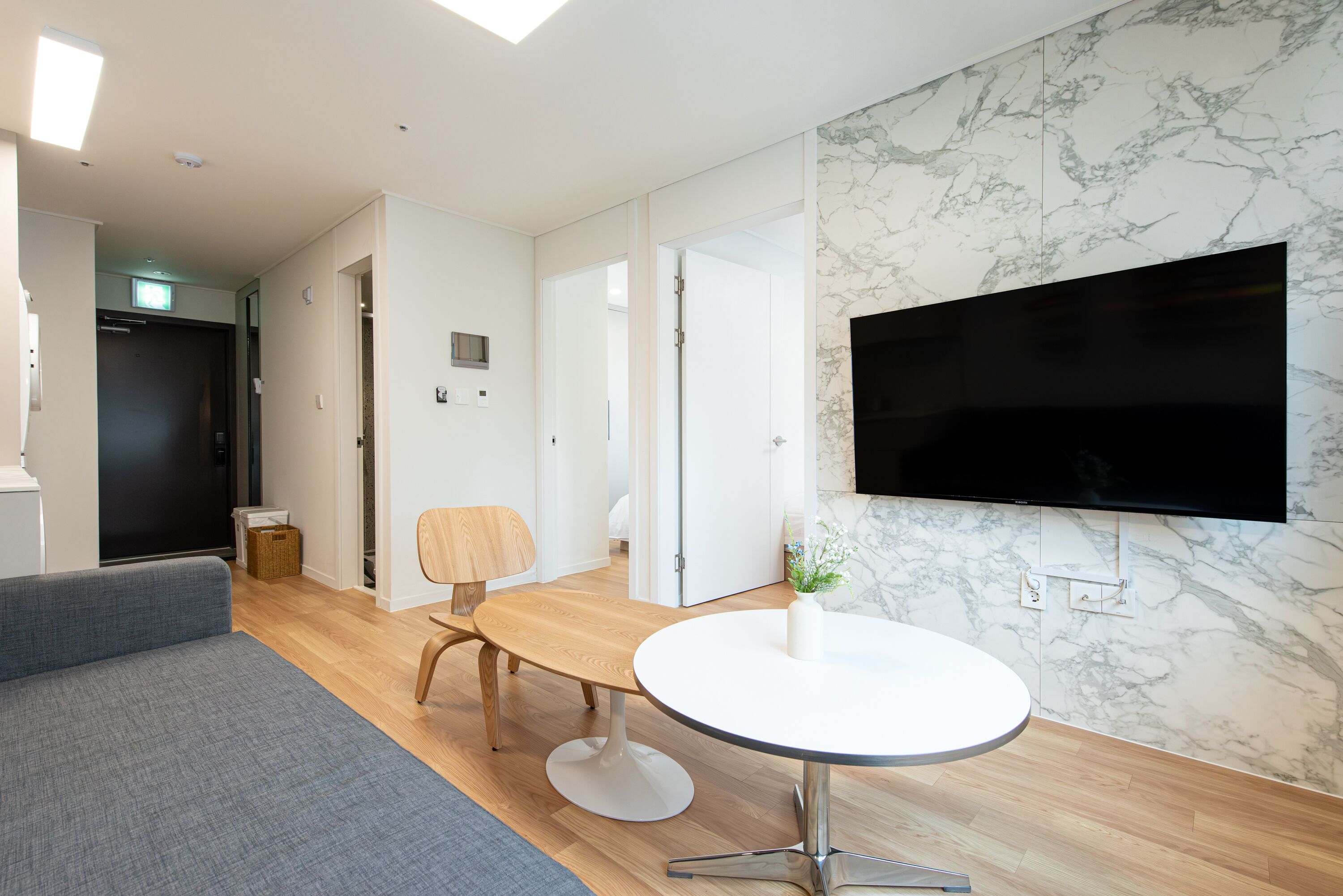 A modern hotel room with a grey sofa, a wooden dining table with a white round table, a flat-screen TV mounted on a marble wall, and a small potted plant.