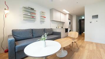 A modern living room with a grey sofa, a round white table, and a wooden dining table. There is a kitchen area with white cabinets and a washing machine.