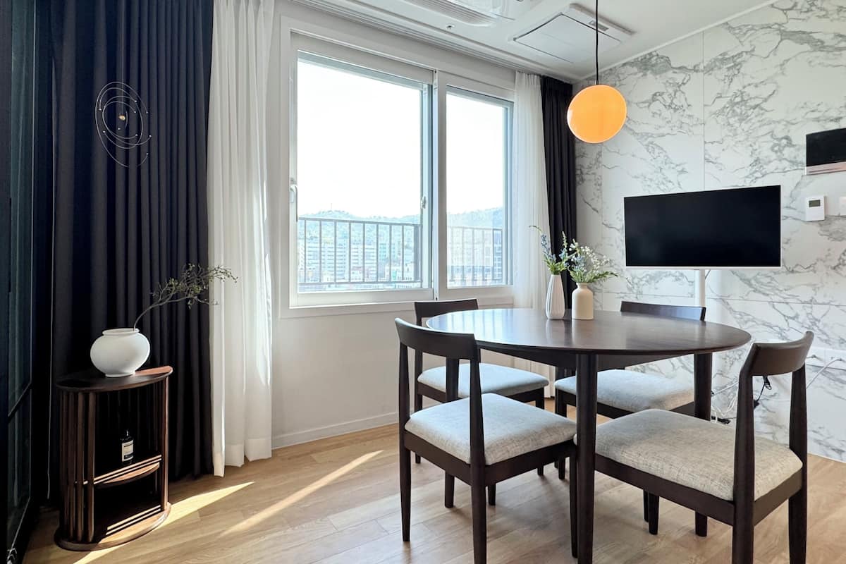 Et moderne spiseområde med et rundt bord og fire stole, et TV ophængt på væggen og et vindue med gardiner.