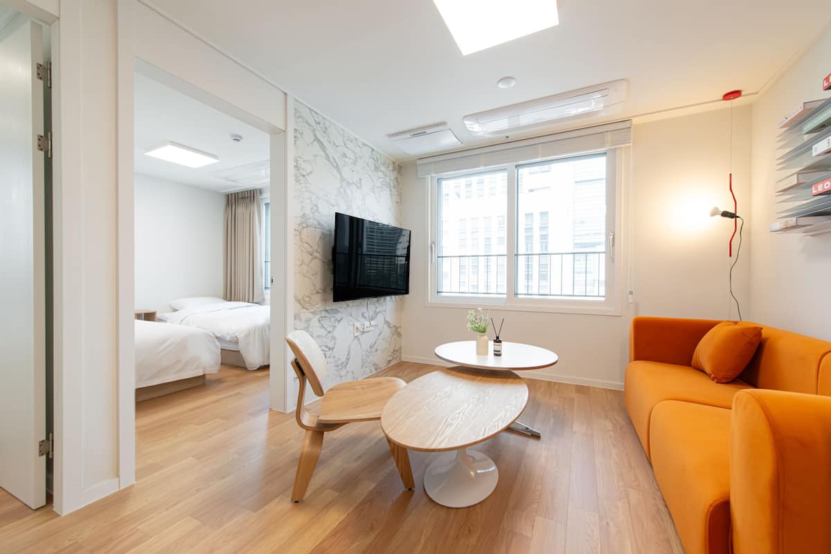 Et moderne hotelværelse med trægulv, et rundt bord, en orange sofa, en seng, et fjernsyn monteret på væggen og et vindue med udsigt over byen.
