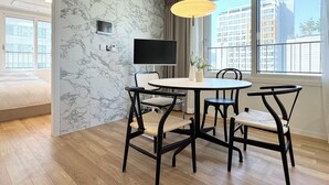 A modern hotel room with a marble wall, a round dining table with chairs, a flat-screen TV, and a large window offering a city view. - GUDO RESIDENCE CHUNGMURO (Seoul)