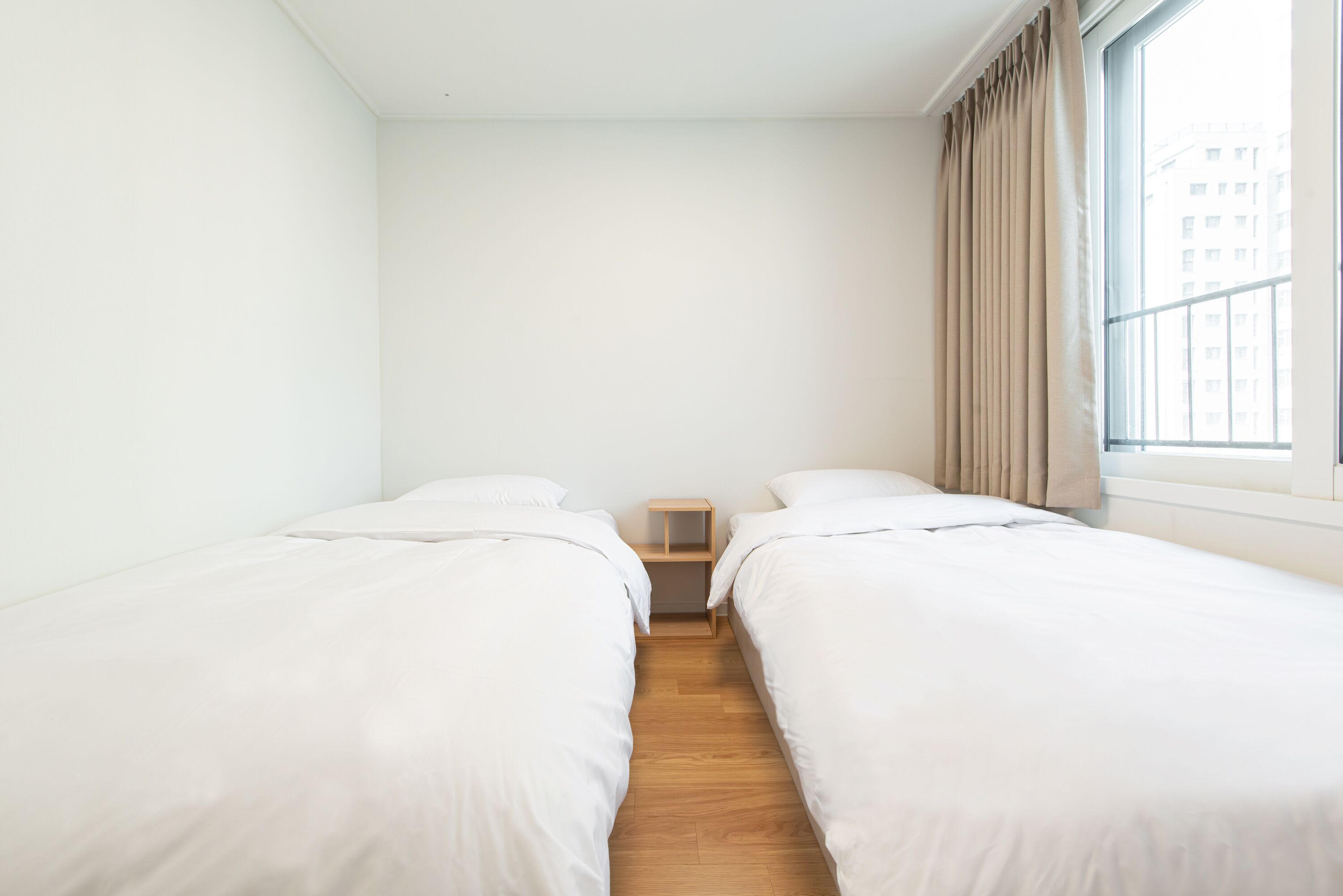 A small, clean room with two single beds, a wooden nightstand, and a window with curtains.