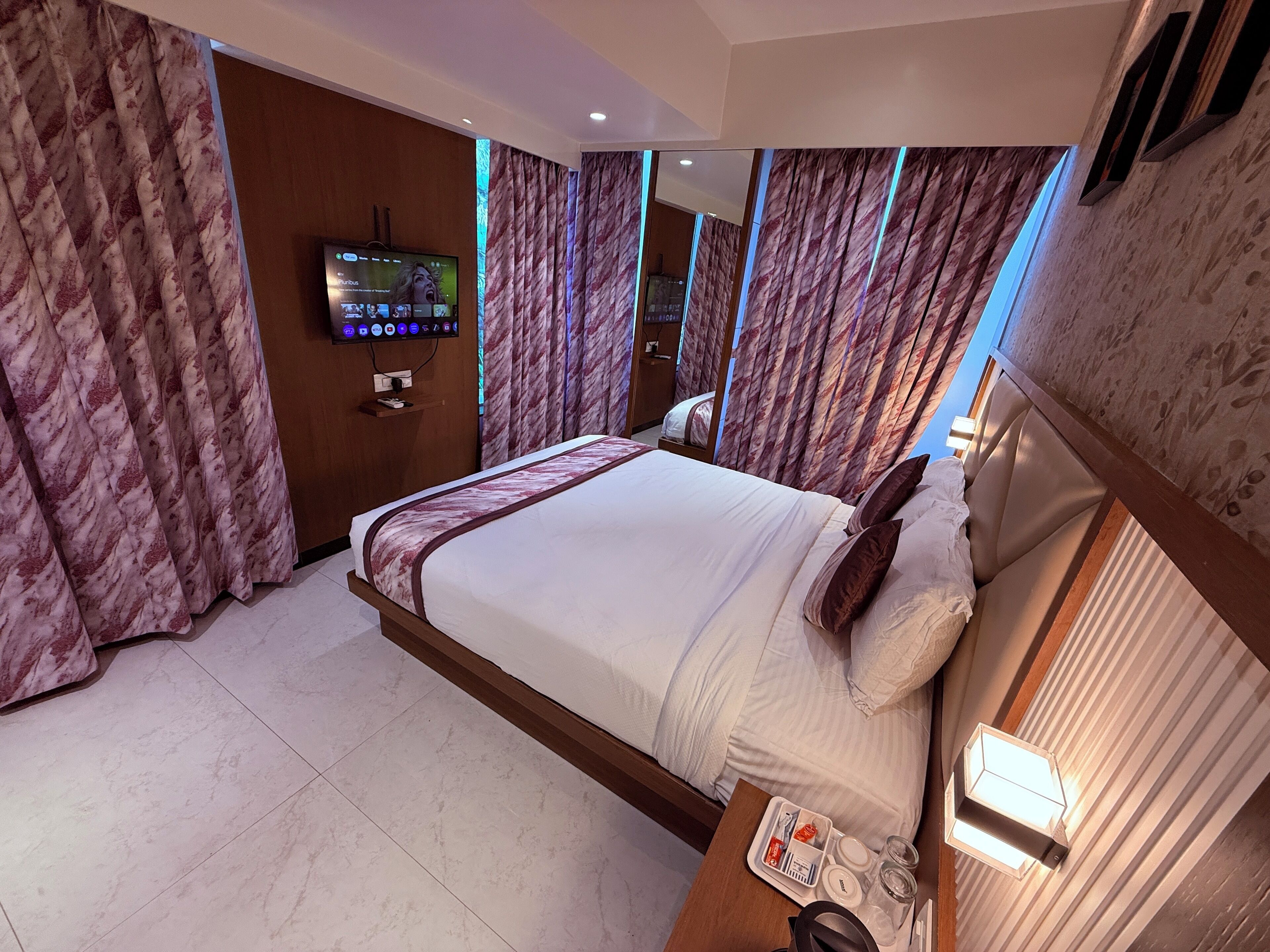 Habitación de hotel con una cama grande, un televisor instalado en la pared y una mesita de noche con una lámpara y un juego de artículos de aseo.