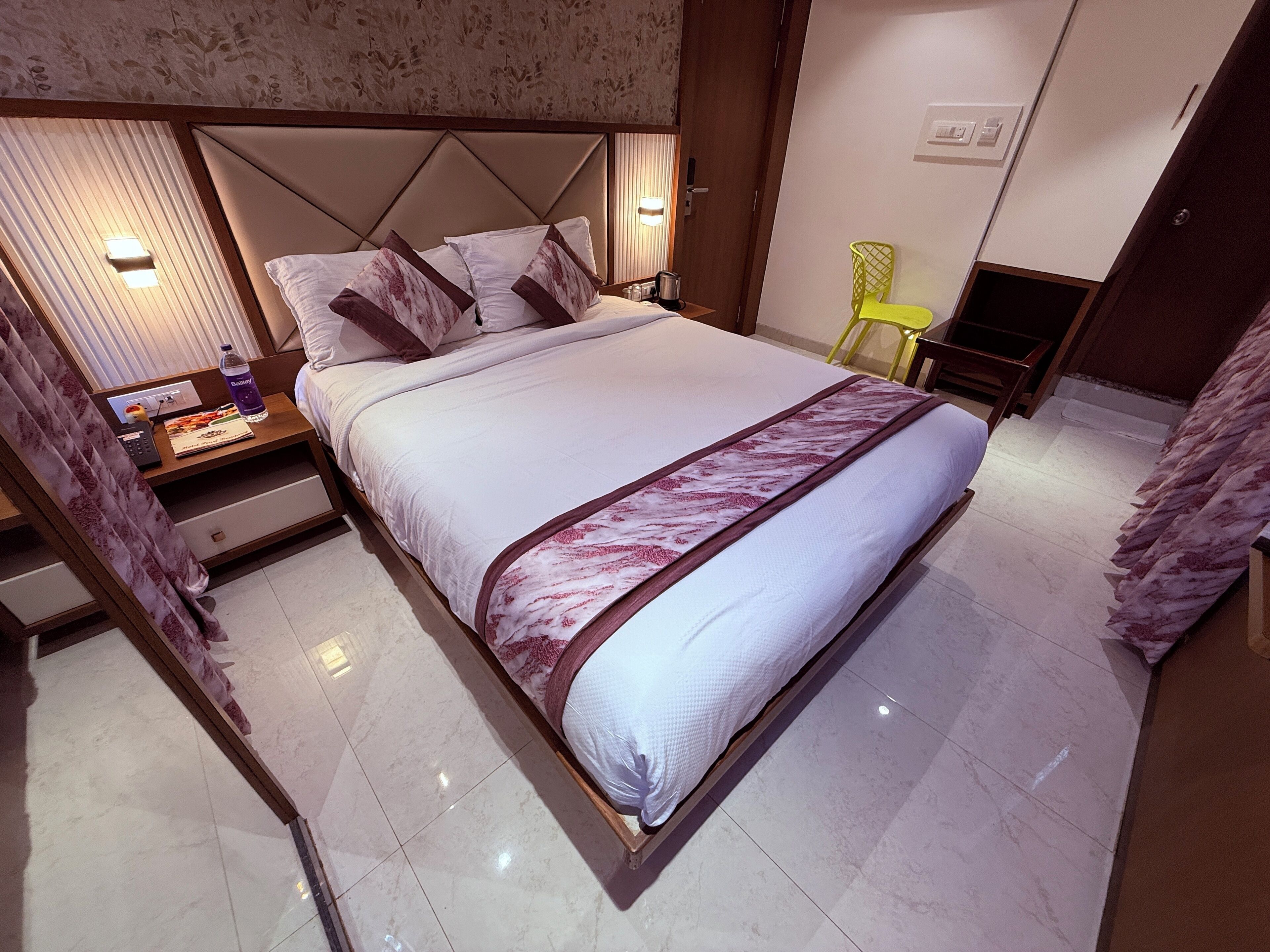 A hotel room with a large bed, a bedside table with a lamp, a TV, and a bathroom area with a shower and toilet.