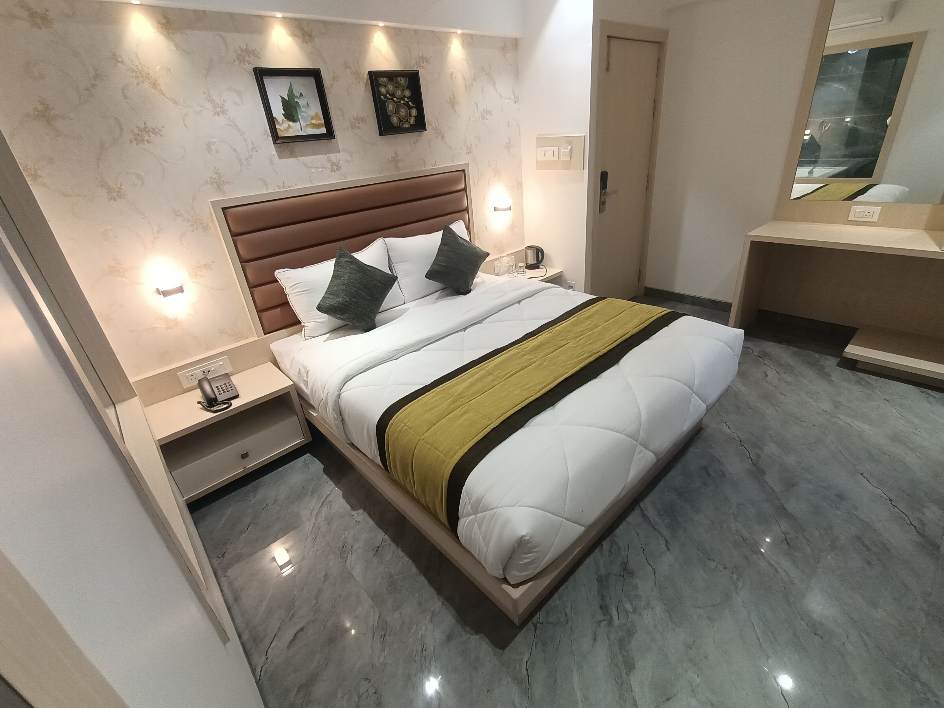 A modern hotel room with a large bed, bedside tables, a nightstand, a mirror, and a wall-mounted light fixture.