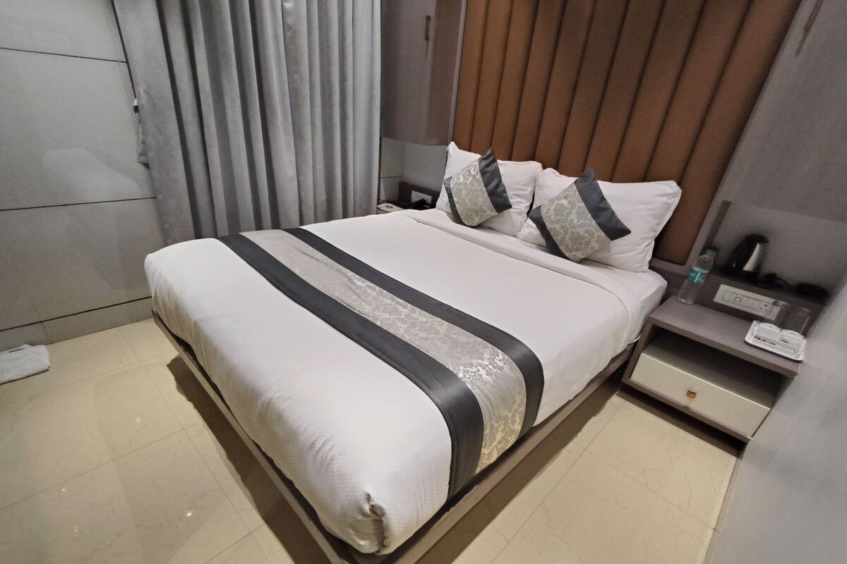 Ein modernes Hotelzimmer mit einem ordentlich bezogenen Bett, einem Nachttisch mit Lampe sowie einem weiteren Nachttisch mit Wasserflasche und Kaffeemaschine.