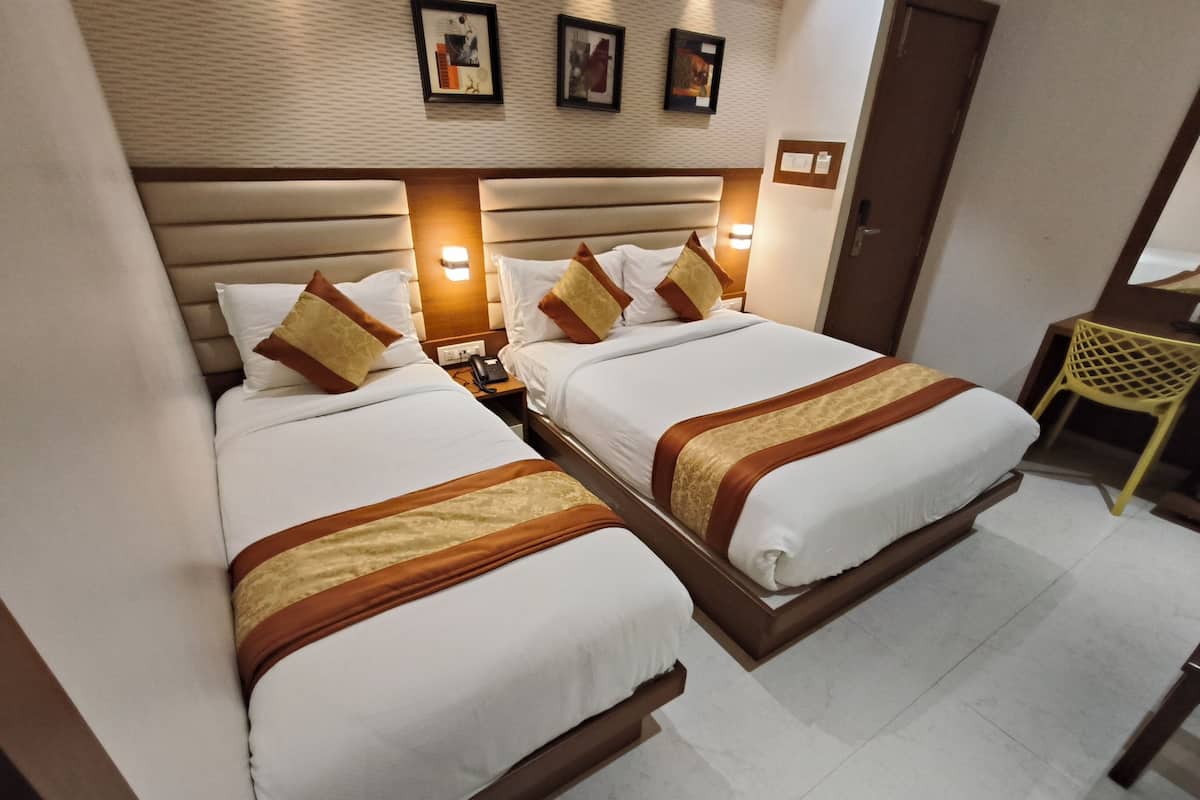 Ein Hotelzimmer mit zwei Betten, jeweils mit weißer Bettwäsche und braunen Zierelementen. Nachttische mit Lampen, ein kleiner Schreibtisch und ein Stuhl gehören zur Einrichtung. Der Raum hat einen Fliesenboden und ein Holz-Kopfteil.