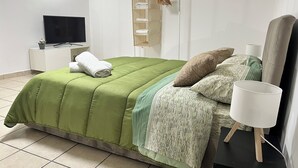 A neatly arranged bedroom with a green bedspread, a television on a white nightstand, a wooden shelf with towels, and a bedside lamp. - Boaters' Home 2, Catania (Catania)
