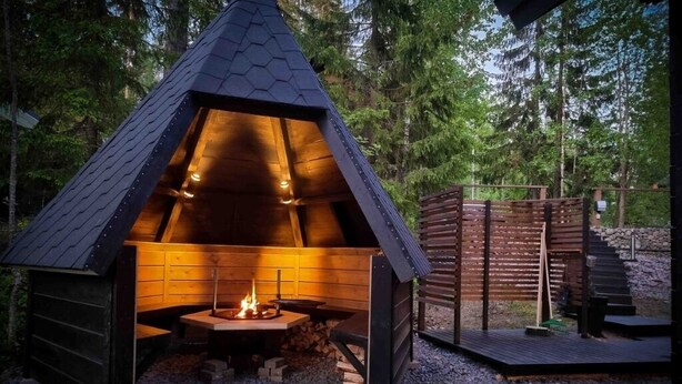 Un espace extérieur accueillant avec un foyer, surmonté d’un toit hexagonal, entouré d’une terrasse en bois et d’un mur en pierre.