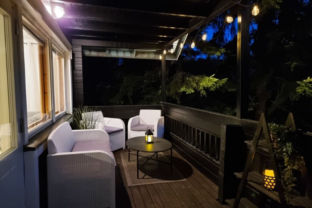 A covered outdoor seating area with wicker furniture and a table, surrounded by windows and a railing.