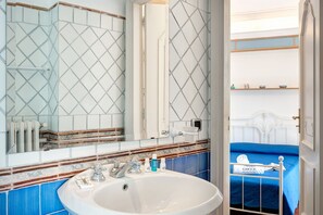 Une salle de bain avec un lavabo blanc, deux robinets et un miroir. On y trouve un lit bleu avec une tête de lit blanche et une serviette qui y est accrochée.
