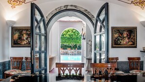 A dining area with a marble table, wooden chairs, and a view of a pool and greenery through an open archway.