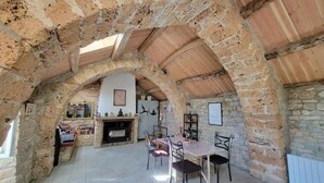 Un intérieur rustique avec des murs en pierre, un plafond en bois, un coin repas avec des chaises et une table, et une cheminée.