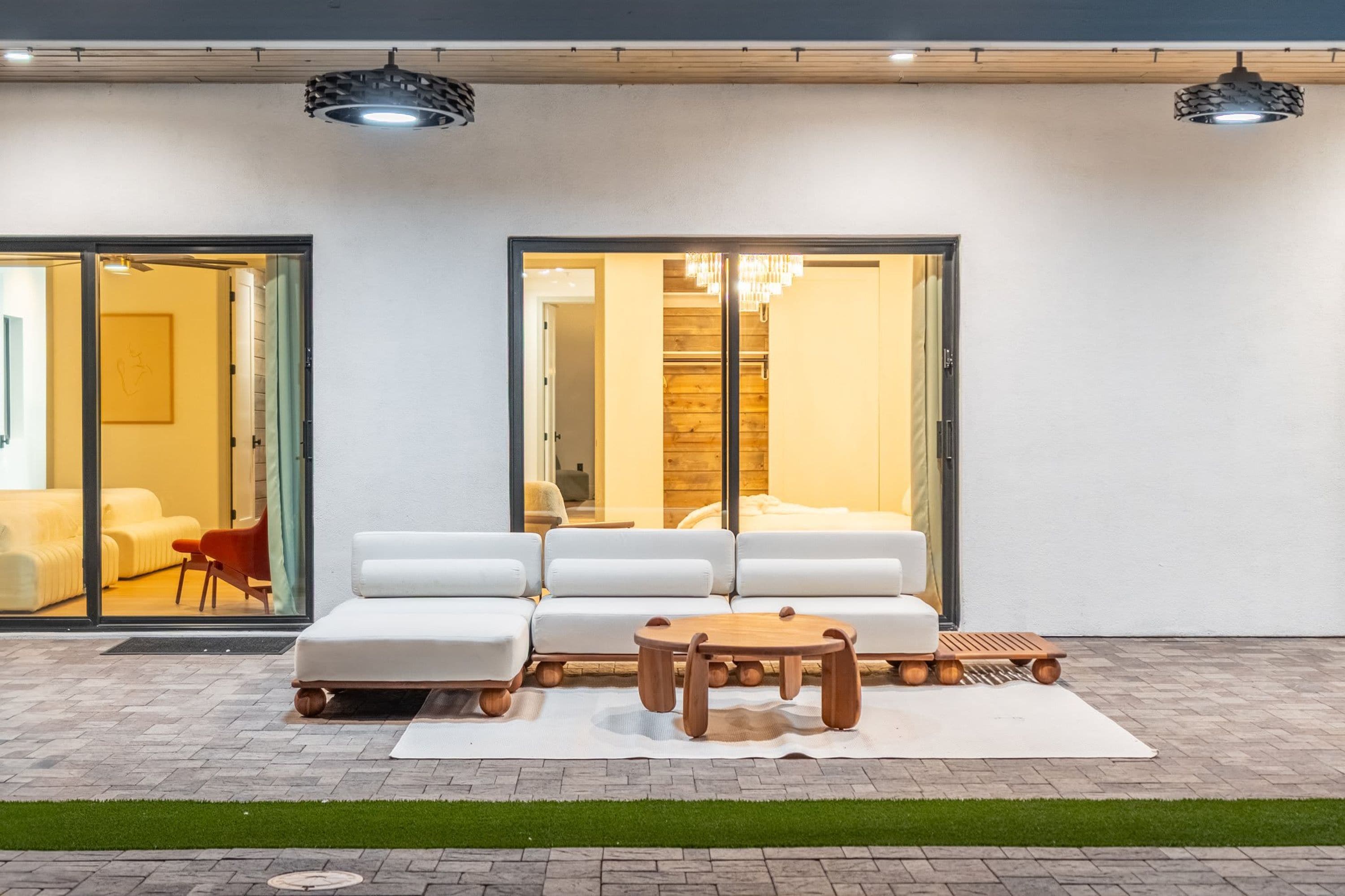 Un área de estar moderna al aire libre con un conjunto de sofás y una mesa de madera, una puerta de cristal que da a un espacio interior, y una araña visible a través de la puerta.