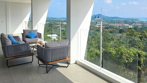 A balcony with two grey upholstered chairs, a wooden table, and a glass railing overlooking a cityscape with greenery and distant hills.