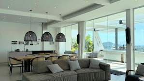 A modern living room with a large sofa, a dining area with a wooden table, and a balcony with a view of the city.